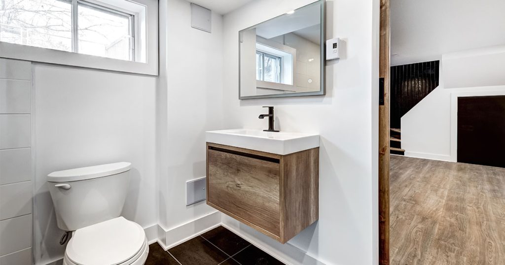 A basement with LVP flooring with a bathroom with black tile flooring and a small modern vanity