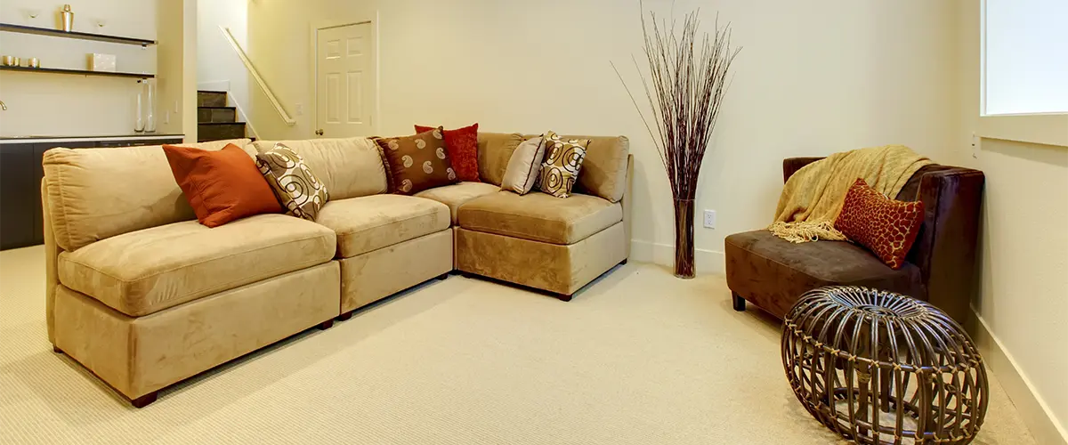 A living room in a basement with a large beige couch and a chair