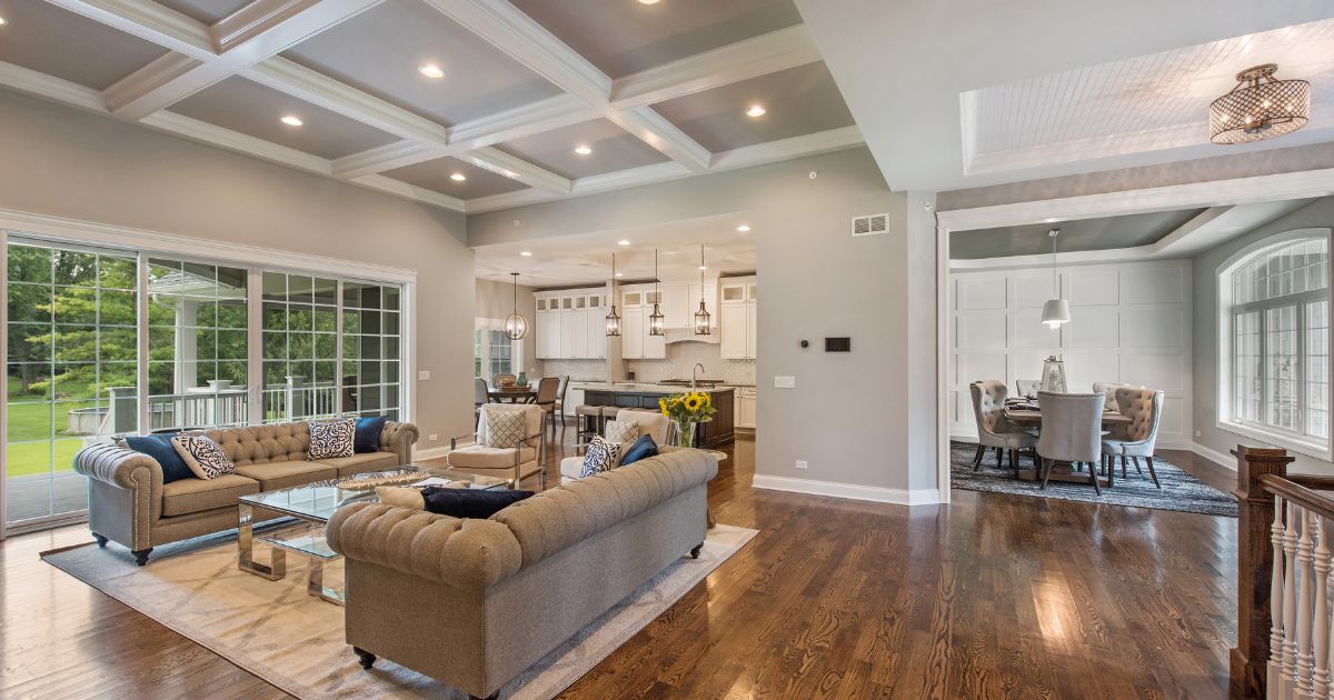 A spacious open-concept living room with coffered ceilings, hardwood floors, and elegant furnishings, seamlessly connecting to a modern kitchen and dining area with large windows and sliding glass doors leading to an outdoor patio.